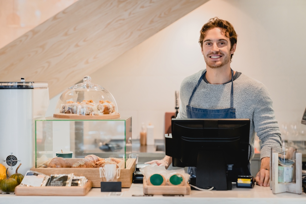 Smiling attractive young waiter standing at cafe counter in small cafeteria. Male cafeteria staff vendor seller selling food and drinks to customers