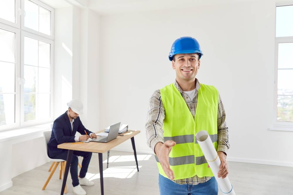 male foreman shaking hand to camera making a deal standing in white impartments holding blueprint with man architect working on laptop